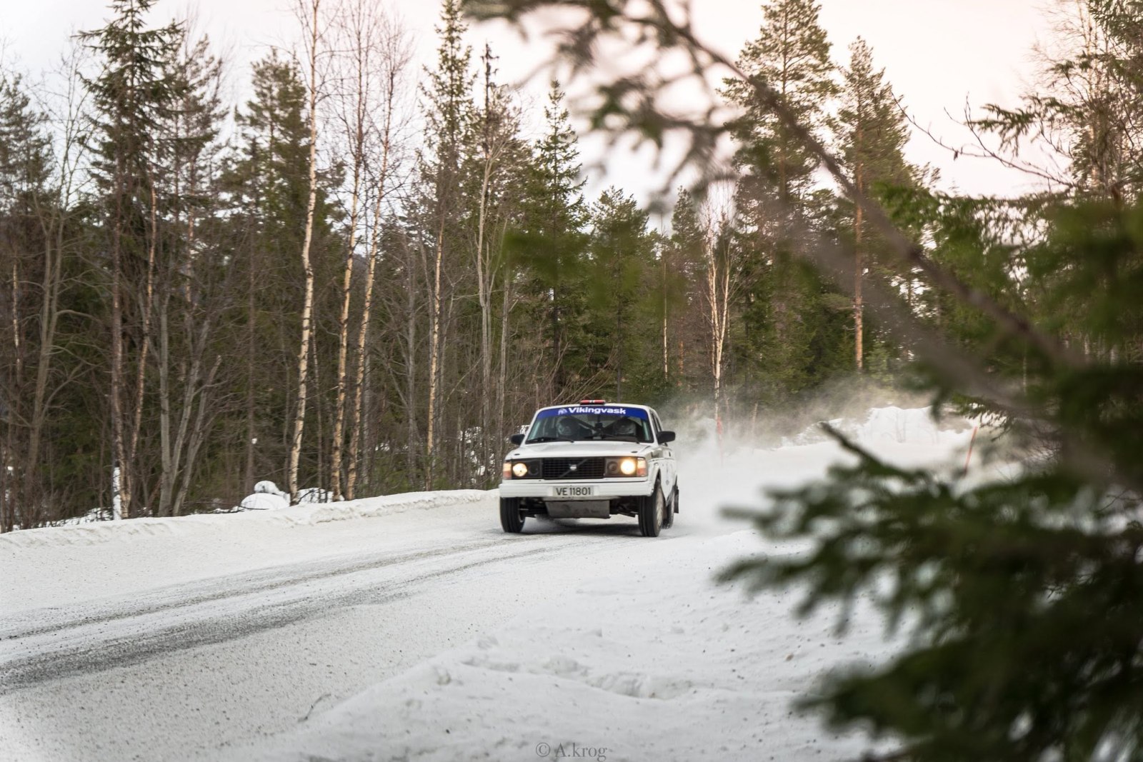 Hvit Volvo på snødekt vei gjennom skogen.